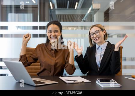 Two happy diverse business women celebrating their success, looking at the camera with arms raised and wide smiles while working together in a modern office environment Stock Photo