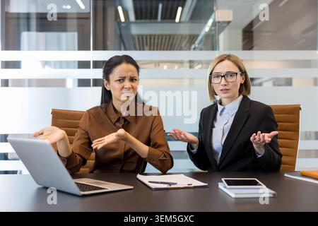 Two diverse businesswomen shrugging their shoulders in an office, expressing confusion, frustration, and misunderstanding during a corporate meeting or discussing a problem Stock Photo