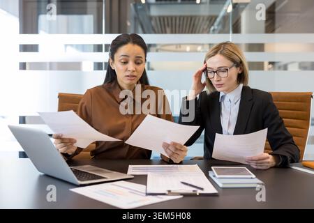 Two business women are reviewing important documents and reports at an office desk, one showing concern while the other exhibits signs of stress and frustration from the task at hand Stock Photo