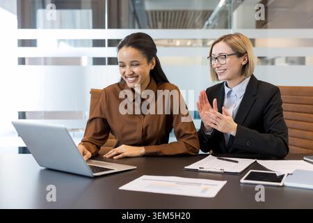 Two diverse businesswomen sitting at a conference table, smiling and looking at a laptop while one woman claps, representing teamwork and achievement in a professional environment Stock Photo