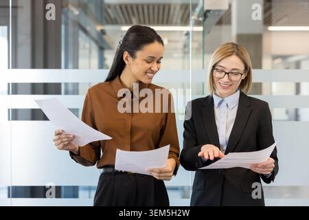Two smiling businesswomen in a modern office collaborate over documents and charts, discussing strategy and analyzing data as they plan a project and celebrate progress together Stock Photo