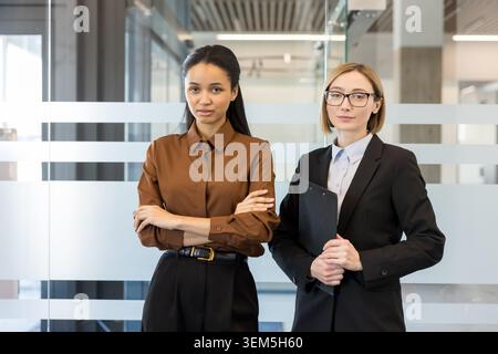 Two confident businesswomen standing in a modern office, one with arms crossed and the other holding a clipboard, conveying diversity, leadership and female empowerment in corporate roles Stock Photo