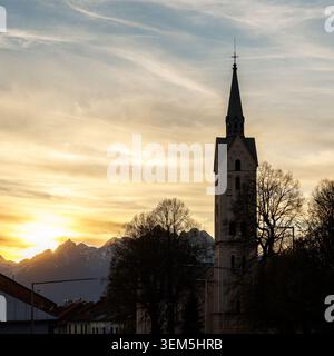 An elegant cathedral spire rises prominently in the foreground with the sharp, snow-dusted peaks of a mountain range glowing under a golden dusk sky. Stock Photo