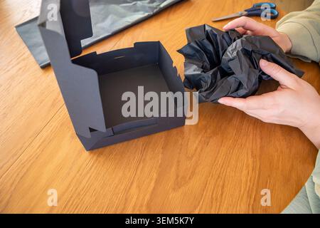 Hands crumpling tissue paper near an open matte black box on a table in natural daylight. Concept: handcrafted gift wrapping Stock Photo