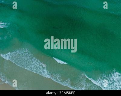 Aerial view of clear turquoise ocean waves rolling onto a sandy beach, creating a calm tropical coastal scene with soft surf and natural textures. Stock Photo