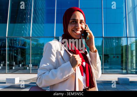 Middle-eastern arab businesswoman wearing hijab strolling in the city - muslim businesswoman wearing hijab using smartphone, smiling in front of a mod Stock Photo
