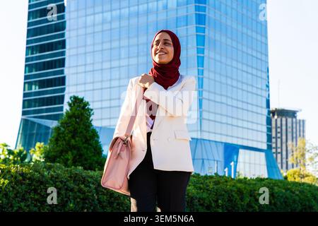 Middle-eastern arab businesswoman wearing hijab strolling in the city - young woman smiling, confidently carrying a bag, walking towards a corporate b Stock Photo