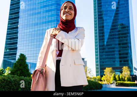 Middle-eastern arab businesswoman wearing hijab strolling in the city - confident businesswoman wearing a hijab and formal blazer smiling with city sk Stock Photo