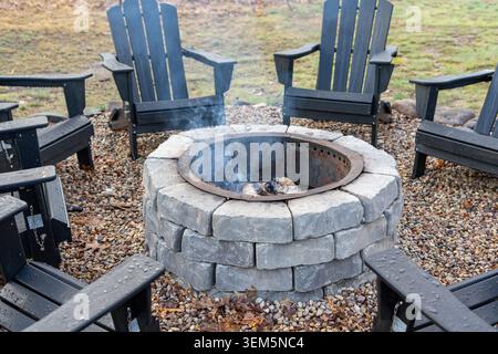 A high-quality medium close-up photograph of a round, custom-built stone brick fire pit with a metal insert. Light smoke rises from a burning wood log Stock Photo