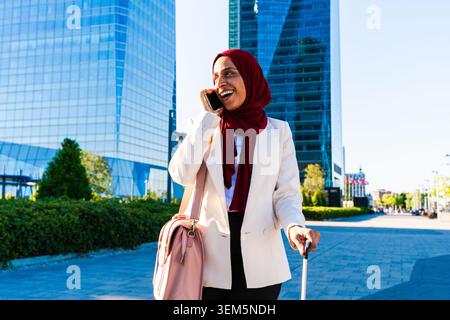 Middle-eastern arab businesswoman wearing hijab strolling in the city - muslim businesswoman wearing hijab walking with luggage and talking on a mobil Stock Photo