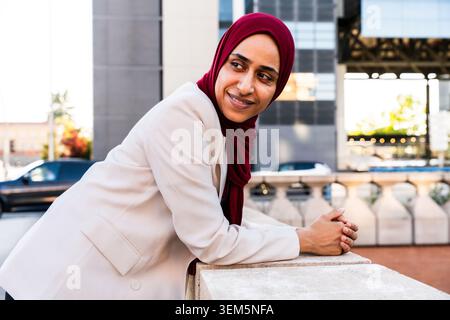 Middle-eastern arab businesswoman wearing hijab strolling in the city - muslim businesswoman wearing a hijab, looking away and smiling in an urban set Stock Photo