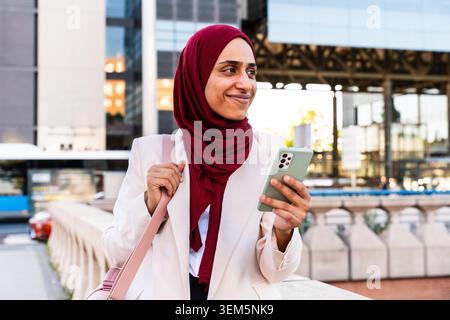 Middle-eastern arab businesswoman wearing hijab strolling in the city - young muslim professional woman in a hijab smiling and checking her phone in a Stock Photo