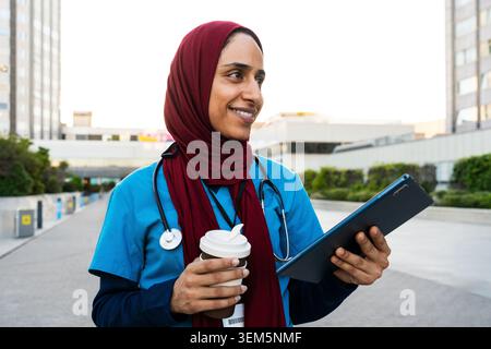 Arabian female doctor wearing a hijab and scrubs is smiling, holding a digital tablet and coffee cup while walking outside a modern hospital building, Stock Photo