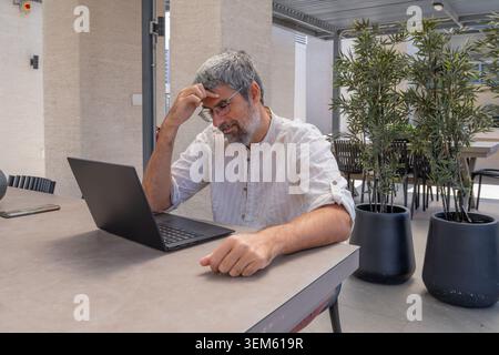 Thoughtful mature man concentrating on his laptop, experiencing stress or deep thought during remote work in an outdoor office setting Stock Photo