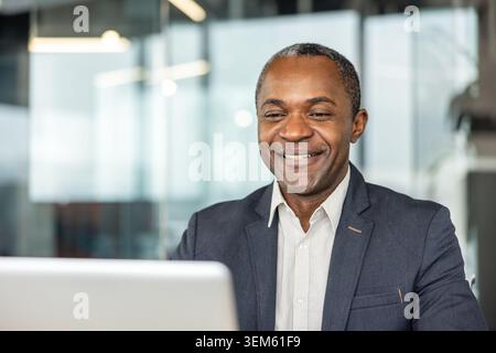 Mature man with a happy expression smiling at the camera, wearing formal attire, and sitting at his desk in a modern office, engaged in a positive work experience Stock Photo