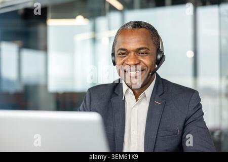 Smiling male call center agent in a suit jacket wearing a headset, confidently assisting clients and working on a laptop in a modern office customer service environment Stock Photo