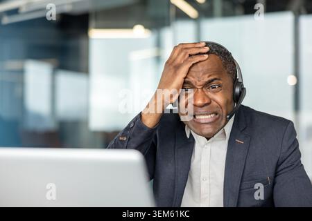 Frustrated mature employee wearing a headset and suit holding his head, experiencing stress and a difficult situation while working on a laptop at his office desk Stock Photo