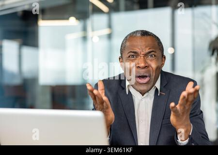 Mature adult businessman expressing intense shock, surprise, and dissatisfaction while raising hands in frustration, reacting to bad news or a problem on a laptop screen in an office setting Stock Photo