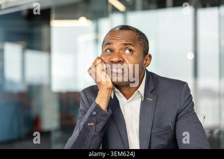 Adult business person working in a modern office, experiencing mental fatigue and boredom, looking upwards with a thinking expression while resting his chin in his hand Stock Photo