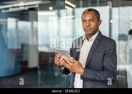 African american businessman using a digital tablet, standing confidently in a contemporary office reflecting professionalism, technology, and leadership in a corporate environment Stock Photo