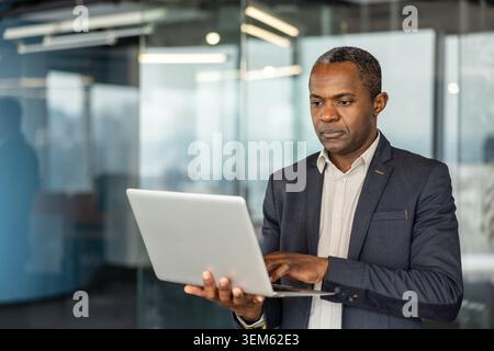 Experienced african entrepreneur focusing on his work while holding a laptop, standing in a contemporary corporate environment, symbolizing dedicated professionalism and technology in business Stock Photo