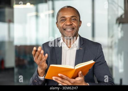 Mature professional man actively engaging in conversation, holding an open notebook, and smiling confidently in a modern office environment, conveying leadership and successful communication Stock Photo