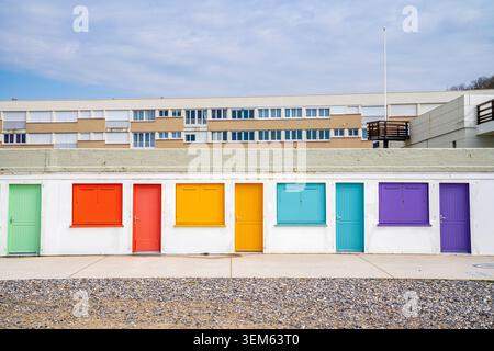 Row of white concrete beach huts with doors and windows in vibrant rainbow colors in Le Treport, France. In the foreground is the pebble beach, while Stock Photo