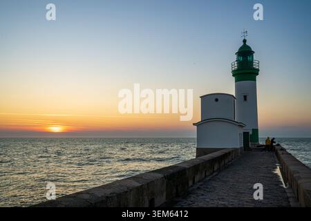 The white lighthouse with a green lantern on the stone pier of Le Treport, France, stands during a scenic sunset over the sea. Two fishermen stand on Stock Photo