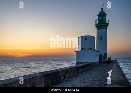 The white lighthouse with a green lantern on the stone pier of Le Treport, France, stands during a scenic sunset over the sea. Two fishermen stand on Stock Photo