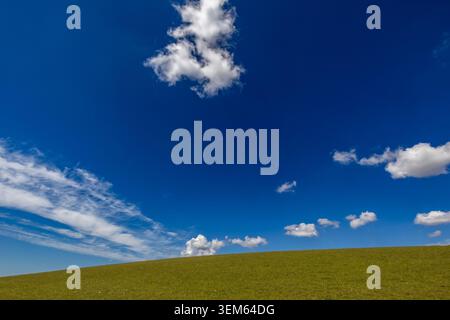 Mixed cumulus and cirrus clouds drift across deep blue sky above a green grass hillside Stock Photo