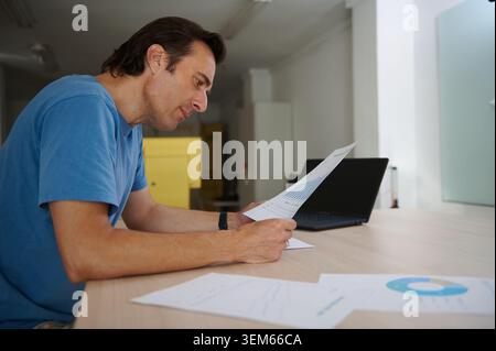 Focused man sitting at a desk and reading a printed sales chart near a laptop in a modern office environment, illustrating financial review, business Stock Photo