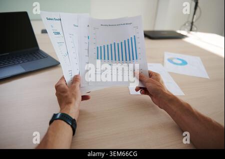 Point of view of hands holding printed sales dashboard reports with charts, graphs and revenue data on a desk with a laptop, representing business ana Stock Photo