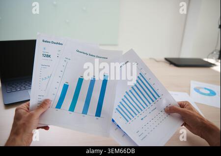 Close-up view of hands holding printed quarterly sales and profit margin reports with charts and dashboard pages on an office desk, representing finan Stock Photo