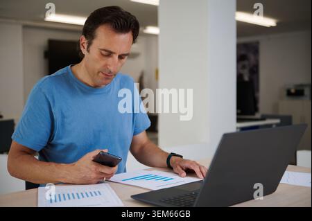 Adult man working with a laptop and smartphone while reviewing printed financial charts on an office desk. The scene is useful for business analytics, Stock Photo