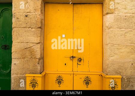 View of a bright yellow wooden door with a padlock set in a traditional limestone wall with floral patterned planters Rabat, Western Region, Malta. Stock Photo