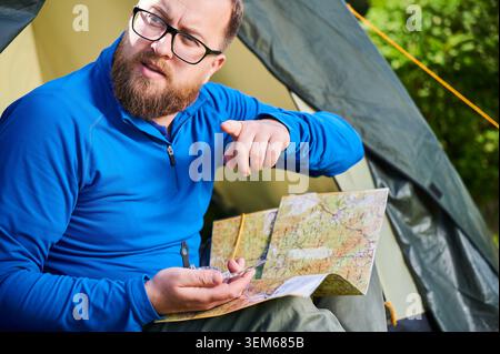 Close up man in glasses, points into distance while holding map and compass. He sits by tent, engaged in navigation, surrounded by greenery and natural landscape. Stock Photo
