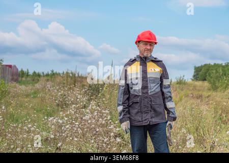 A male engineer in a red hard hat and work jacket stands on a grassy field. land inspection, worker in protective gear, occupational safety. Stock Photo