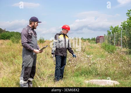 Workers inspect the state of utility networks on a field plot near a concrete hatch. technical maintenance, engineering survey, infrastructure control Stock Photo