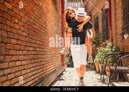 Happy interracial couple having fun piggyback ride in Tbilisi old city narrow street.Cheerful young man giving girlfriend piggyback ride.Couple laughi Stock Photo