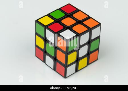 A Rubik's Cube sits on a white table. The colors are bright and arranged in a mixed pattern. Hands may try to solve it. Stock Photo