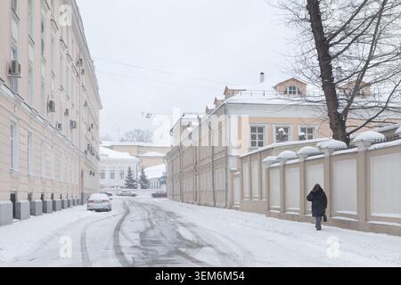 View of a snowy street lined with historic pastel-colored buildings and a person walking along a snow-covered sidewalk in Moscow, Moscow, Russia. Stock Photo