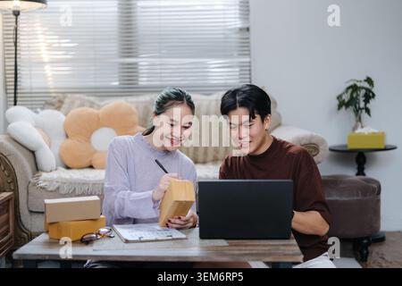 Smiling Asian couple writing on parcel box while using laptop at home with cardboard boxes. E-commerce, small business, online seller and startup life Stock Photo