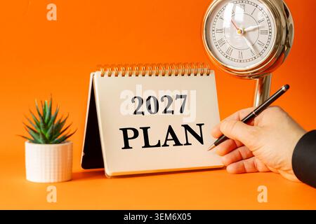 Hand holding pen writing in desk calendar with text 2027 PLAN next to vintage clock on bright orange background Stock Photo