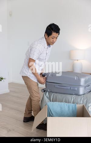 Middle-aged Asian man bending over handling gray suitcase on bed beside open box with blue towel Stock Photo