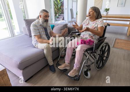 Man sitting on sofa talking to senior woman in manual wheelchair at home, holding pink bouquet Stock Photo
