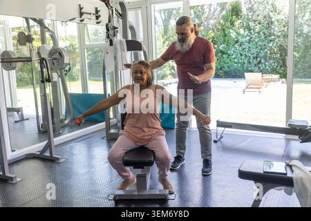 Senior African American woman in pink doing lateral raises on bench with weights, trainer guiding Stock Photo