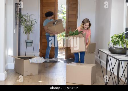 Couple carrying packed moving boxes and potted plants into home foyer, wooden door open, copy space. Cardboard, relocation, greenery, foliage, entrywa Stock Photo