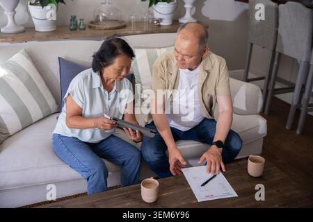 Asian couple sitting on sofa in living room wearing casual attire reviewing bills with tablet Stock Photo
