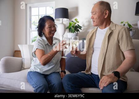 Asian couple sitting on light-colored sofa clinking stemmed white wine glasses in living room Stock Photo
