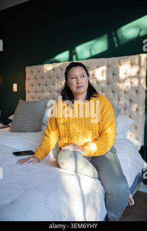 Mature woman sitting on bed edge by tufted headboard in yellow sweater, smartphone resting nearby Stock Photo